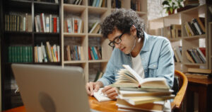 Student sat at a library desk writing in a notebook, with research books and a laptop also on the desk