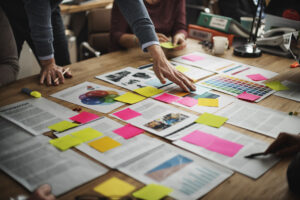 Paper spread acorss a desk labelled with sticky notes, being reviewed by a group