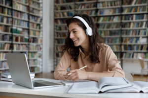 Student sat at a library desk writing in a notebook, with research books and a laptop also on the desk